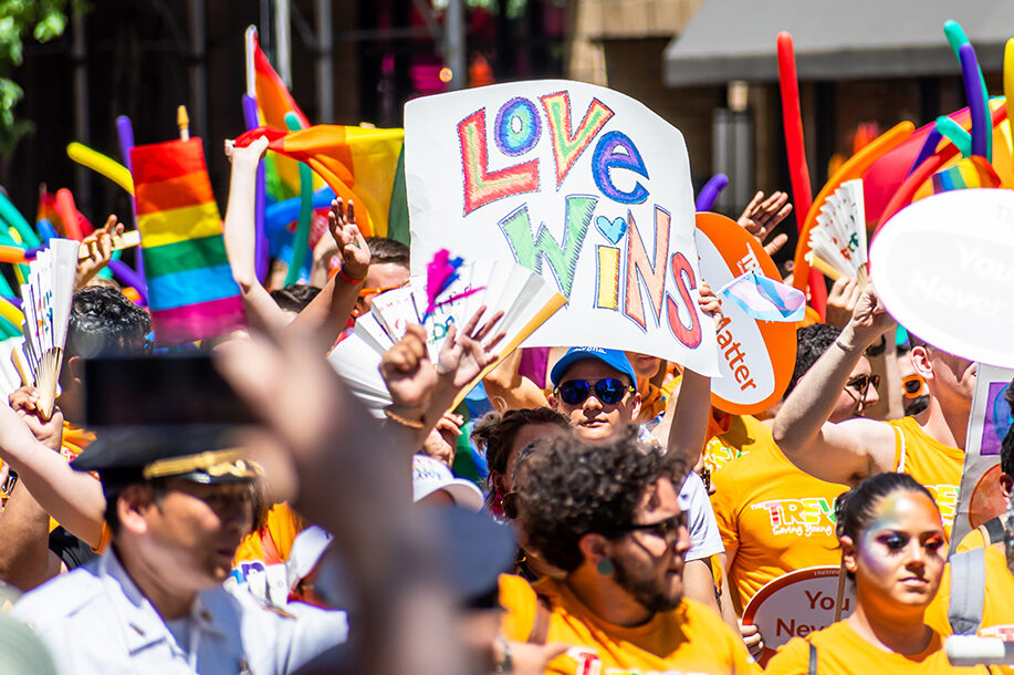 Person holding a Love Wins sign amongst thousands of people wearing colorful costumes attending the World Pride March in NYC for LGBTQ+ rights.